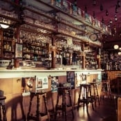 Bar aux murs de bouteilles et tabourets hauts dans une lumière chaude, salle Speakeasy chez Tempus Fugit à Beauchamp