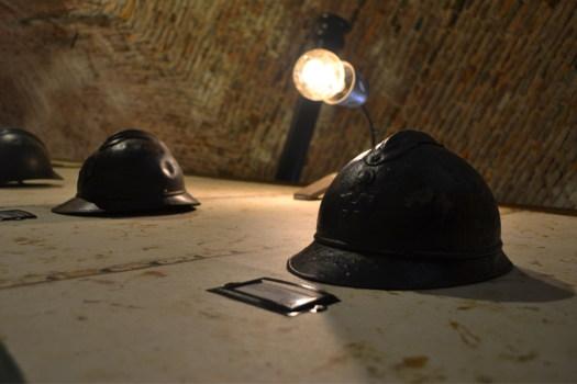 Casques militaires sombres posés sur une table sous une lampe, salle 1916 chez La Clé du Bastion à Belfort