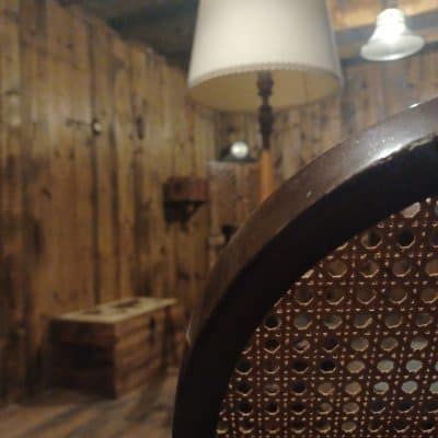 Dossier de chaise cannée devant un mur de planches et une lampe à abat-jour, salle L'héritage de tante Suzanne, Zone 72