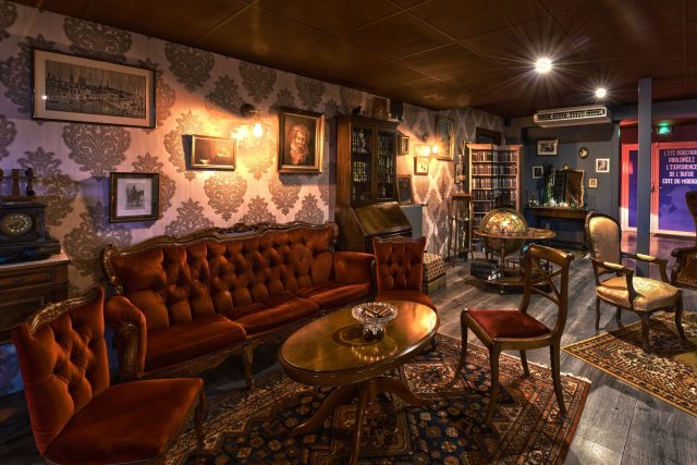 Intérieur d'une bibliothèque vintage avec canapé en velours roux, tableaux et globe terrestre chez Escape Hunt Nantes