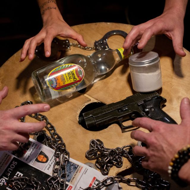 Mains de joueurs sur un plateau avec bouteille, pistolet, menottes et chaînes, salle Le Cartel chez The Box Escape Games