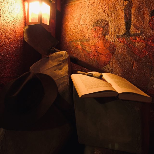 Décor souterrain aux murs ocre, livre ouvert éclairé par une lanterne rougeâtre dans Le Temple