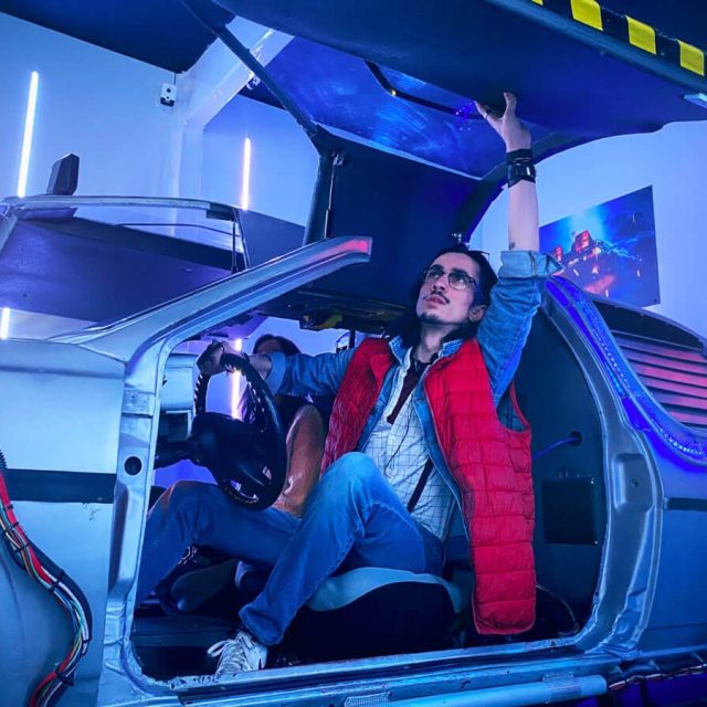 Joueur au gilet rouge assis dans une voiture à portes papillon, décor bleu, salle Retour dans le temps chez Freeing à Saint-Maximin