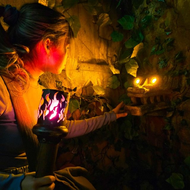 Joueuse approchant une torche à flamme rouge d'un mur de pierre couvert de plantes, salle Tomb Hunter chez Freeing à Saint-Maximin