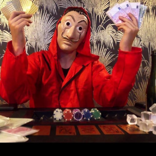 Figurant en costume rouge et masque doré devant décor à motifs, jetons de poker sur table