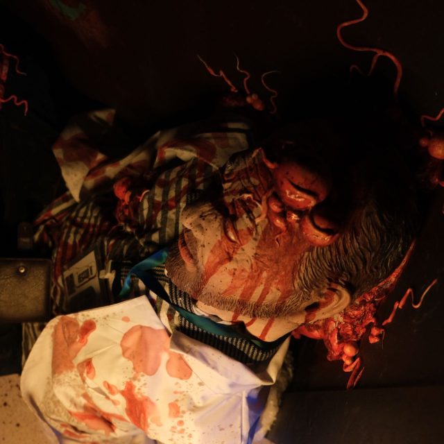 Personnage au tablier ensanglanté et tentacules rouges sur fond sombre, salle Le virus chez Rashomon Escape