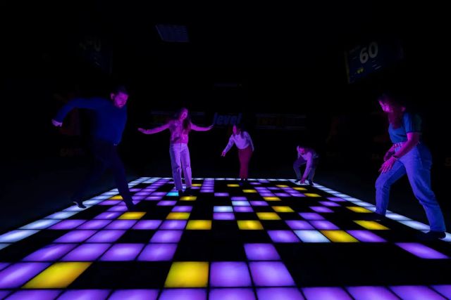 Joueurs sur un sol de dalles lumineuses violettes et jaunes dans le noir, salle Level UP chez Le Voyage Heure à Besançon