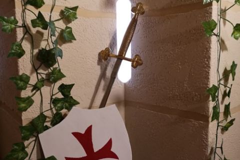 Bouclier blanc à croix rouge et épées contre un mur couvert de lierre, salle Exploration au bastion chez Enigm'Antic à Périgueux
