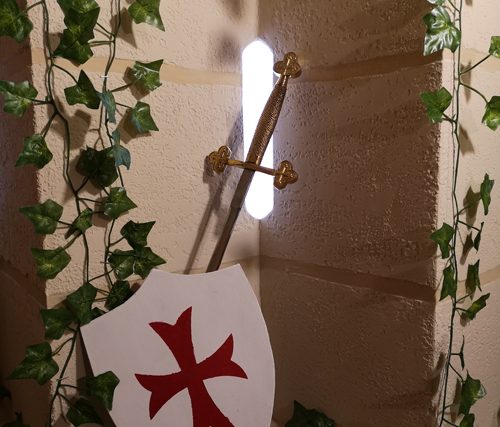 Bouclier blanc à croix rouge et épées contre un mur couvert de lierre, salle Exploration au bastion chez Enigm'Antic à Périgueux