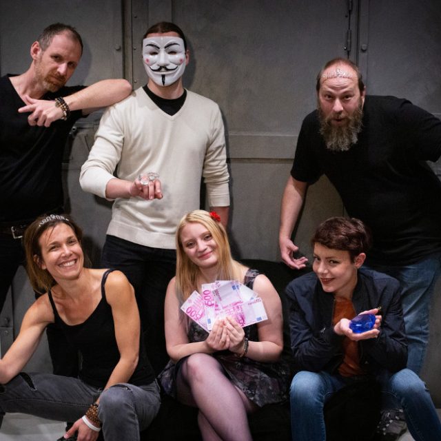 Groupe de joueurs souriants dans une salle The Box Escape Games, l'un portant un masque blanc