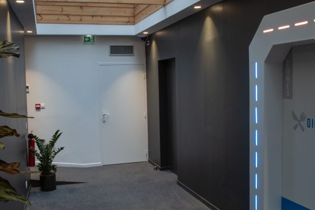 Couloir moderne aux murs gris, plante verte et porte de sas futuriste lumineuse, salle Cybercity chez Just Escape à Lille