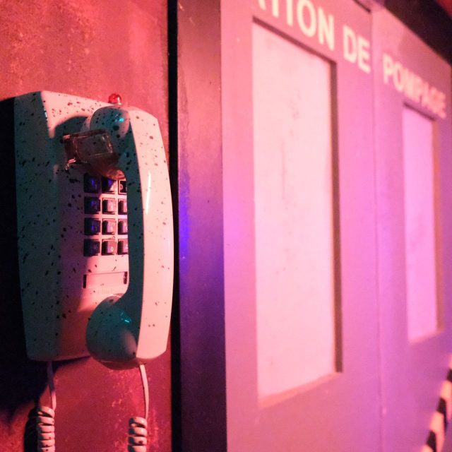 Téléphone mural gris près de portes éclairées en rose et rouge, salle Le virus chez Rashomon Escape