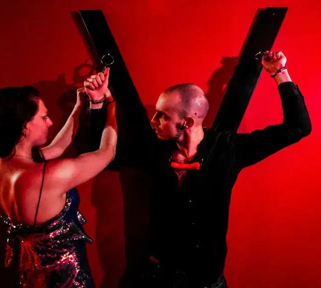 Deux joueurs menottés à une croix en bois sur fond rouge, salle Blackmail chez Evade