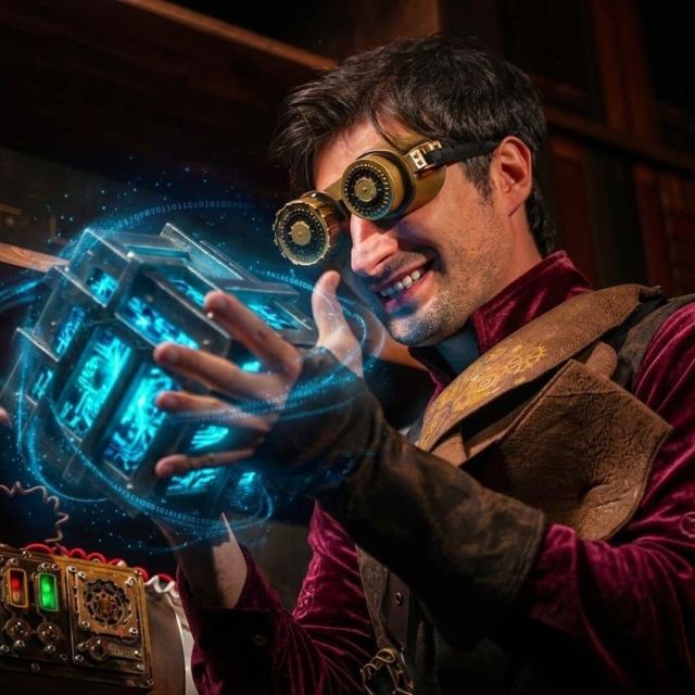 Homme aux lunettes à engrenages brandissant un cube bleu éclatant, salle Steampunk chez BlackOut Room à Nice