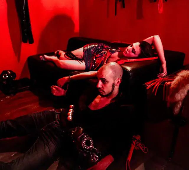 Homme assis par terre et femme allongée derrière lui, décor rouge de la salle Blackmail