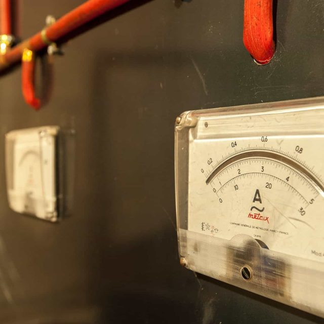 Ampèremètre vintage et câblage électrique rouge sur panneau gris dans Le Blockhaus : Double enjeu