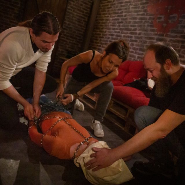 Joueurs penchés sur une personne en combinaison orange attachée au sol, salle Le Cartel chez The Box Escape Games