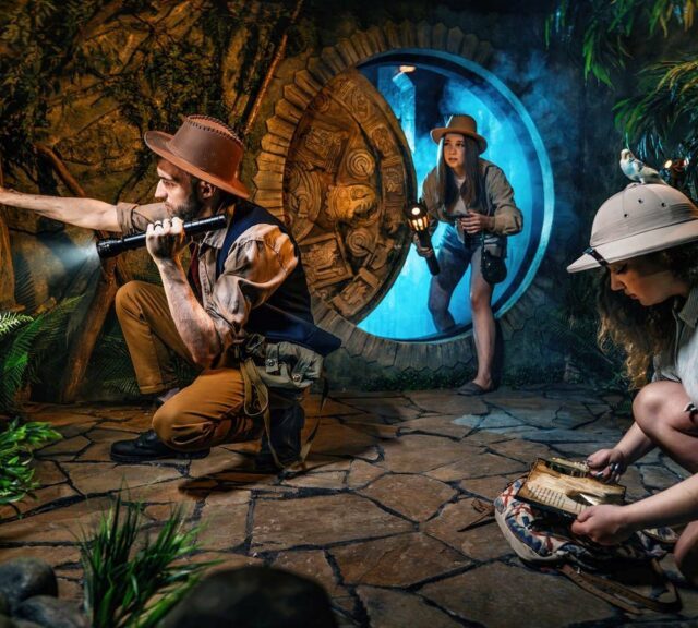 Trois explorateurs en chapeau devant un disque de pierre gravé dans une jungle, salle Indiana, One O'Clock