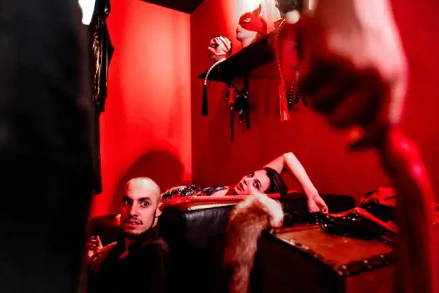 Deux joueurs allongés dans une pièce rouge, une main tenant un fouet au premier plan, salle Blackmail