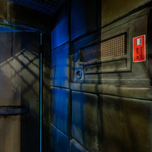 Couloir d'asile aux murs métalliques gris, éclairage bleu froid, panneau d'alerte rouge