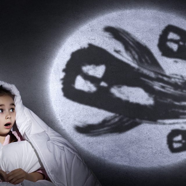 Enfant sous draps blanc face à des ombres spectrales de créatures ailées sur mur illuminé