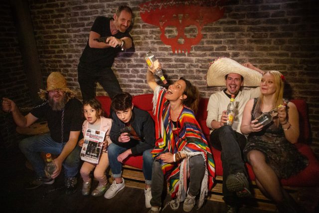 Joueurs en ponchos et sombreros sur une banquette rouge, un homme buvant, salle Le Cartel chez The Box Escape Games à Metz