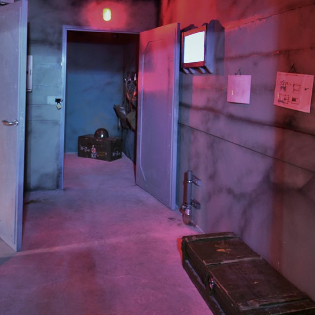 Intérieur de bunker aux murs rose-rouge, portes métalliques et objets de mission chez Team Break Terville