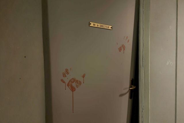 Porte grise marquée d'empreintes de mains rouges sous une plaque de directeur, salle L'Aile maudite
