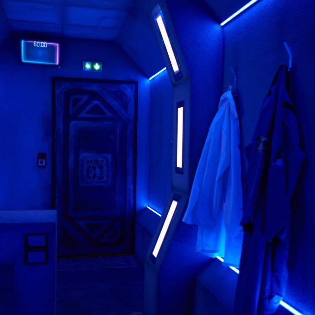 Couloir de vaisseau éclairé de néons bleus avec blouses accrochées, salle Le portal chez Escape One à Pontault-Combault