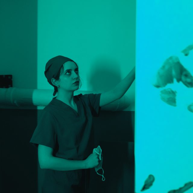 Infirmière en blouse verte devant un mur turquoise marqué d'empreintes de mains, salle La morgue, Rashomon Escape