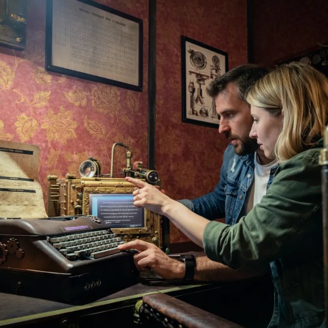 Homme et femme penchés sur une machine à écrire reliée à un écran, salle Le retour de Sherlock Holmes chez Freeing à Osny