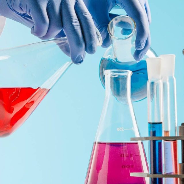 Mains gantées versant liquide rouge dans fiole, flacons colorés en arrière-plan scientifique