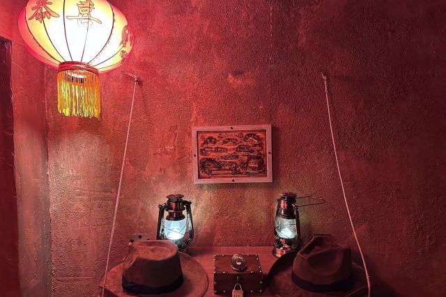Lampion rouge, carte encadrée, lanternes et chapeaux d'aventurier sur un mur rouge, salle Le temple sacré chez Fun Room à Lons