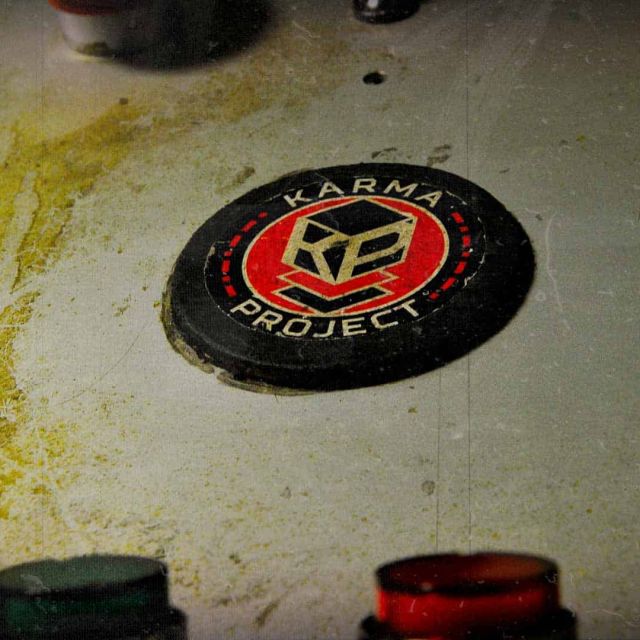 Bouton rond au logo Karma Project posé sur un plan de travail taché de jaune, salle Karma Project, Prizoners