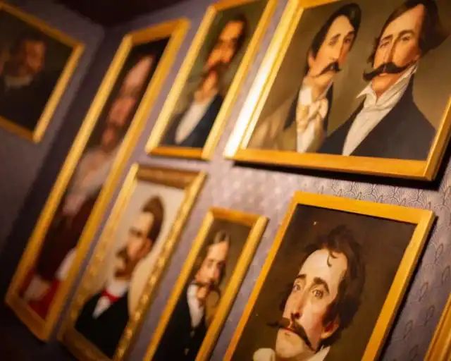 Portraits d'hommes moustachus aux cadres dorés accrochés sur un mur bleu, salle La fortune du fondateur chez Gaillard Académie