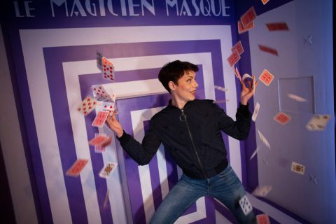 Joueuse entourée de cartes qui volent devant un mur à rayures violettes et blanches, salle La Grande Illusion, The Box Escape Games