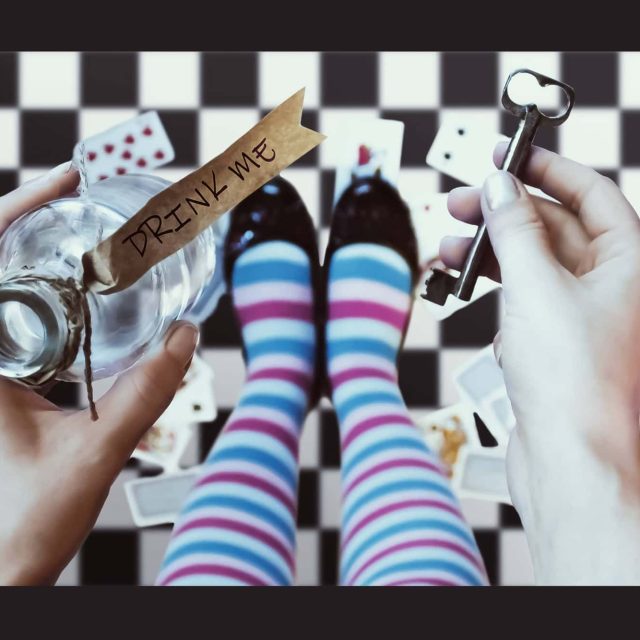 Deux mains tiennent une clé et une fiole sur un damier noir et blanc, ambiance Alice au pays des merveilles chez Le Chapelier fou