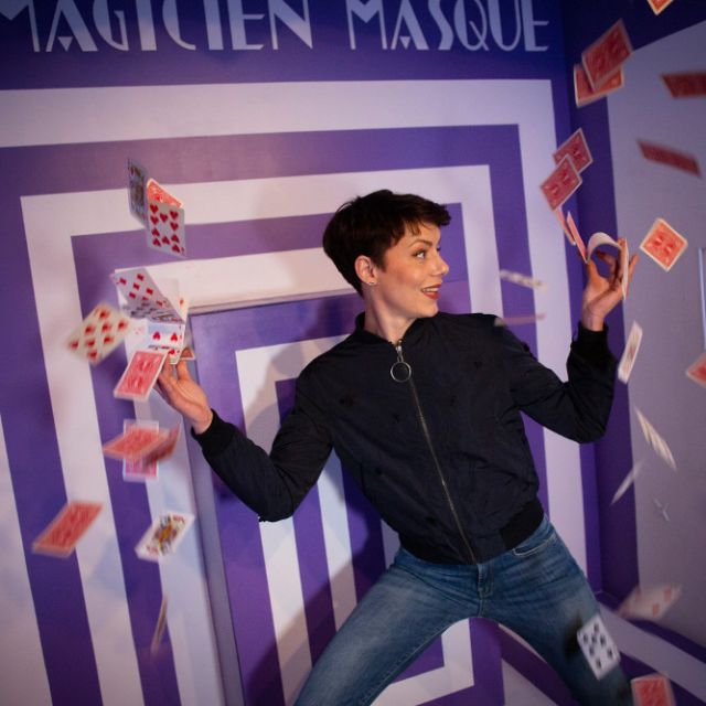 Femme en veste noire lançant des cartes à jouer dans un décor de couloir violet en perspective, La Grande Illusion à Metz
