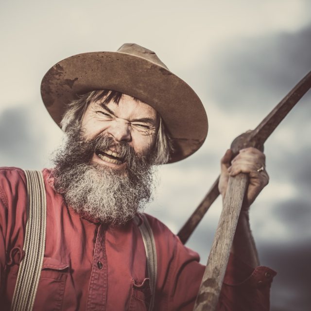 Homme barbu en chemise rouge et chapeau de cowboy tenant un fusil sur fond de ciel nuageux