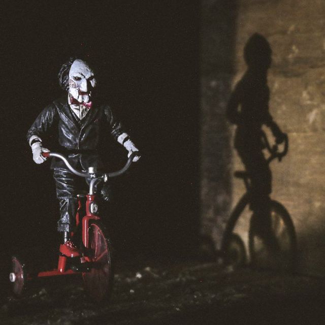 Poupée au visage blanc et joues rouges sur un tricycle rouge, ombre au mur, salle Sacrifice, One O'Clock