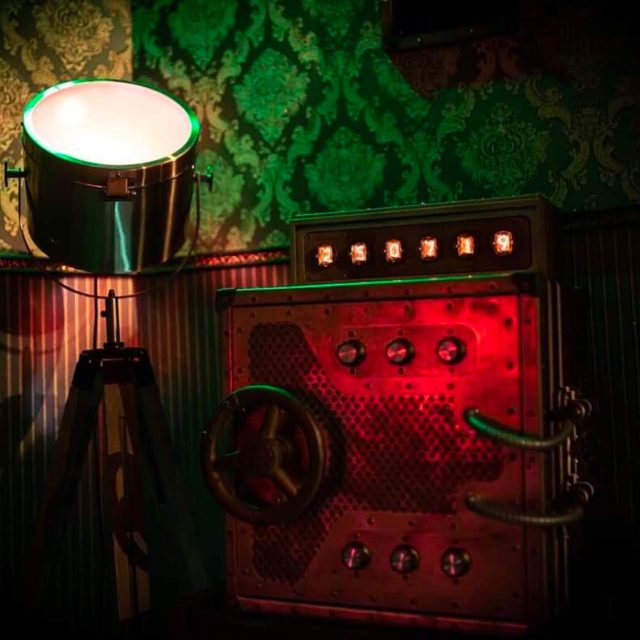 Projecteur sur trépied, boîtier rouge à voyants et papier peint vert à motifs, salle Sherlock Holmes chez Freeing à Saint-Maximin