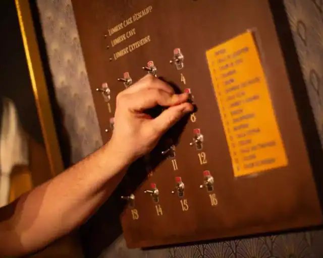 Main manœuvrant des interrupteurs sur un tableau de bois, liste jaune à côté, salle La fortune du fondateur chez Gaillard Académie