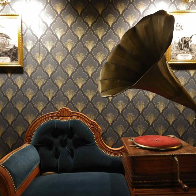 Gramophone à pavillon doré près d'un fauteuil bleu devant un papier peint art déco, salle Sur les traces du magicien à Potelle