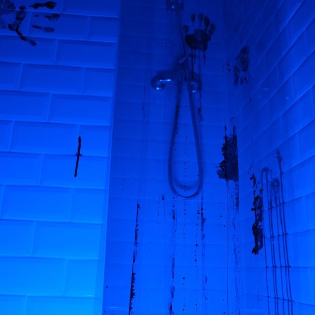 Mur carrelé bleu avec tuyau de douche et traces sombres, salle La morgue chez Rashomon Escape à Paris