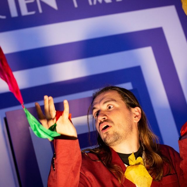 Magicien aux longs cheveux en veste rouge tirant des foulards colorés, salle La Grande Illusion, The Box Escape Games à Metz