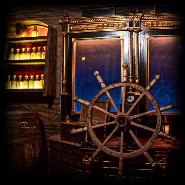 Barre à roue de navire dans une cabine de bois aux étagères éclairées, salle Le trésor de Barbe Noire à Reims
