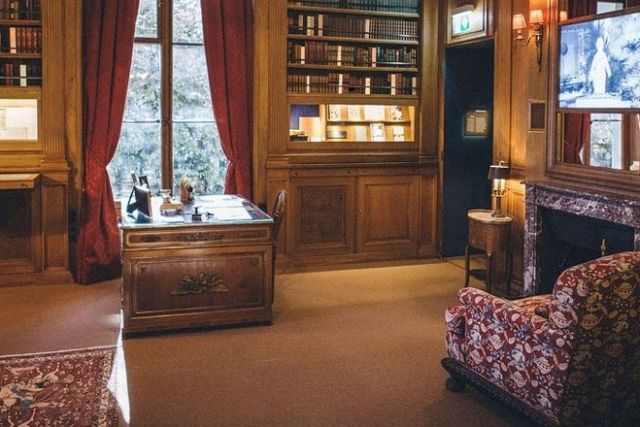 Bureau de bois avec bibliothèque, cheminée, rideaux rouges et fauteuil fleuri, salle Le bureau d'oncle Glenn chez Easy rider park
