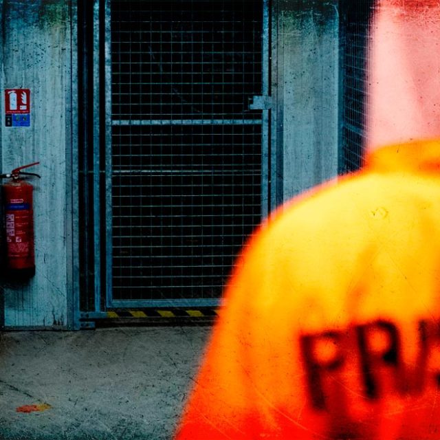 Couloir de prison avec gilet orange marqué "PRISONER", murs bleus et briques, extincteur