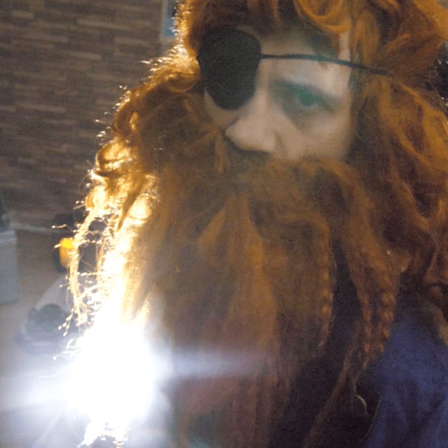 Personnage avec barbe rousse et accessoires fantaisistes dans un décor intérieur chez Escape Game Aventure à Puteaux