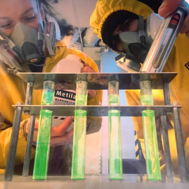 Deux joueurs en combinaisons jaunes manipulent des éprouvettes de liquide vert fluorescent